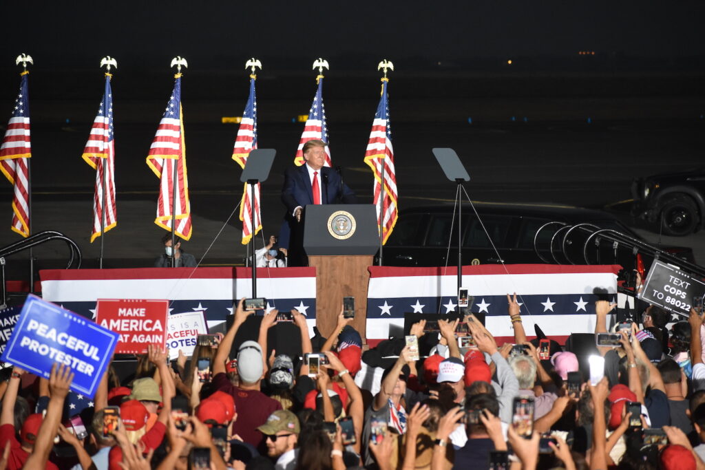 El presidente Donald Trump habla desde un podio. En primer plano, aparece una gran multitud de seguidores con carteles y teléfonos móviles. Varias banderas estadounidenses se muestran detrás de Trump.