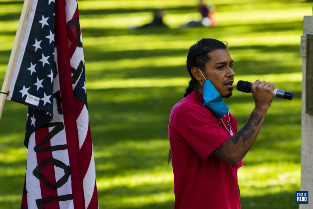 un hombre con una camisa roja y una máscara alrededor de una oreja sostiene un micrófono. Está parado al lado de una bandera estadounidense y hay césped detrás de él.