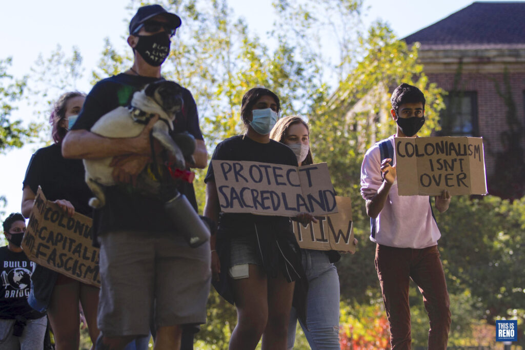 Un grupo de personas marcha. Una persona sostiene un perro. La gente sostiene carteles en los que se lee: "Protege la tierra sagrada" y "El colonialismo no ha terminado".
