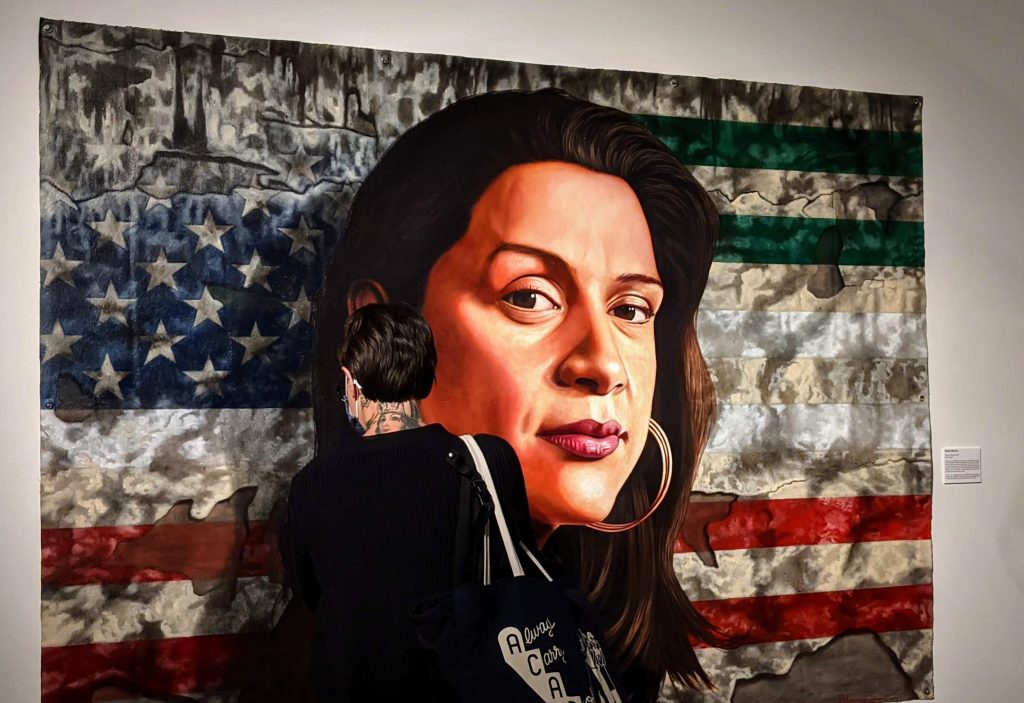 Persona mira de cerca un cuadro con el rostro de una mujer frente a las banderas de Estados Unidos y México