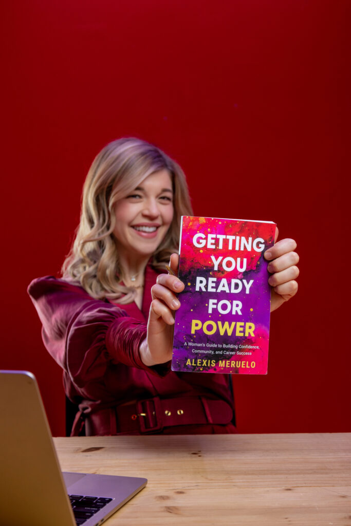 Una mujer rubia sentada en un escritorio sosteniendo un libro que dice: "Preparándote para el poder."
