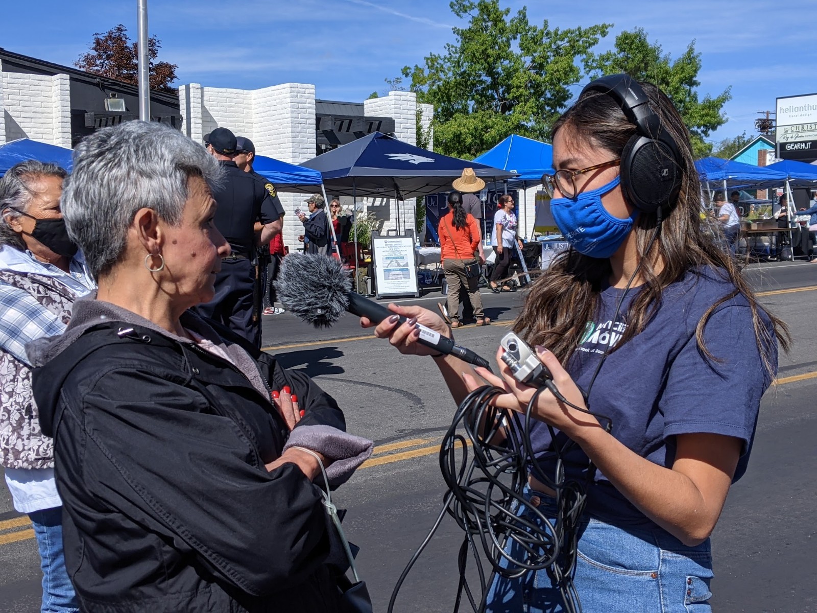 Una reportera con cubreboca, micrófono, grabadora y audífonos, entrevista a una señora en medio de un festival callejero.