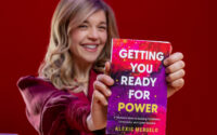 Una mujer rubia sentada en un escritorio sosteniendo un libro que dice: "Preparándote para el poder."